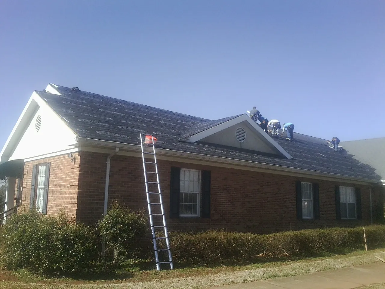Crew tearing shingles off a brick ranch roof with a ladder set — Emergency Roof Repair work by our crew in Ama