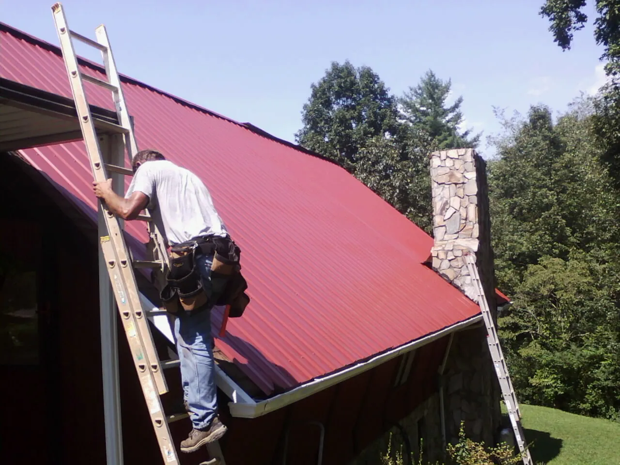 Roofer on a ladder beside a red metal roof and stone chimney — Soffit & Fascia Repair by Roofing Brothers in Ama